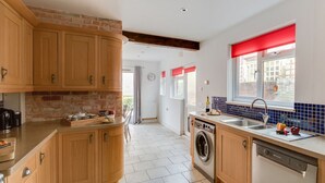 A kitchen with wooden cabinets, a built-in washing machine, a dishwasher, and a window with red curtains. - Sandsfoot Rest (Weymouth)