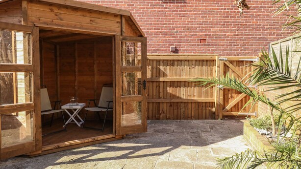 A wooden outdoor seating area with a table and chairs, a wooden fence, and a brick building in the background. - Sandsfoot Rest (Weymouth)