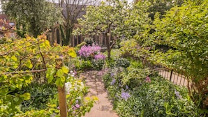 Un sentier de jardin bordé de différentes plantes et arbres, menant à un bâtiment à la façade en briques et aux fenêtres en arc de cercle.