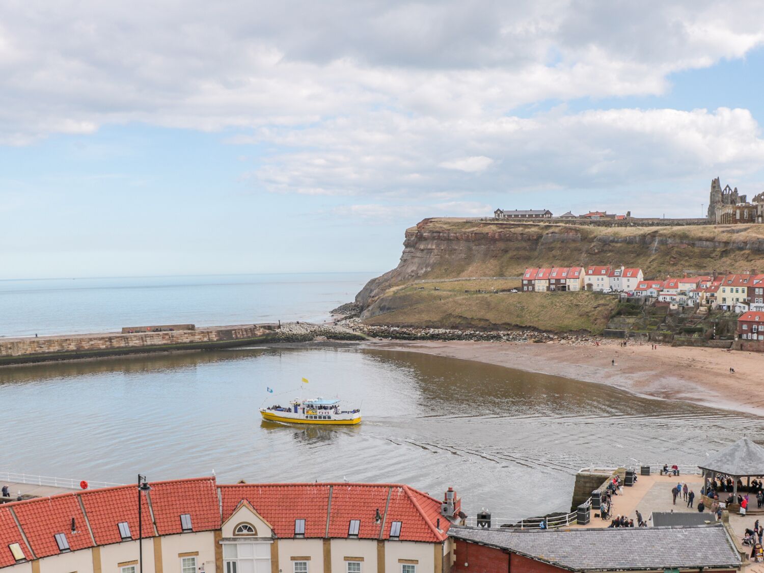 Une ville côtière avec un château historique perché sur une falaise, un port où se trouve un bateau, et une promenade animée.