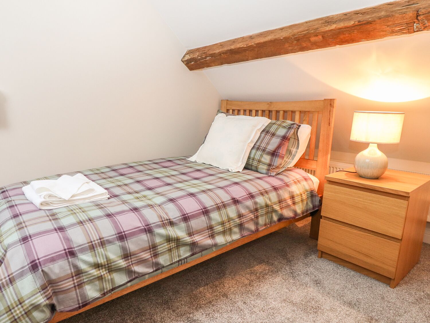Una cama individual con colcha escocesa, cabecera de madera, mesita de noche con lámpara y un cajón pequeño.