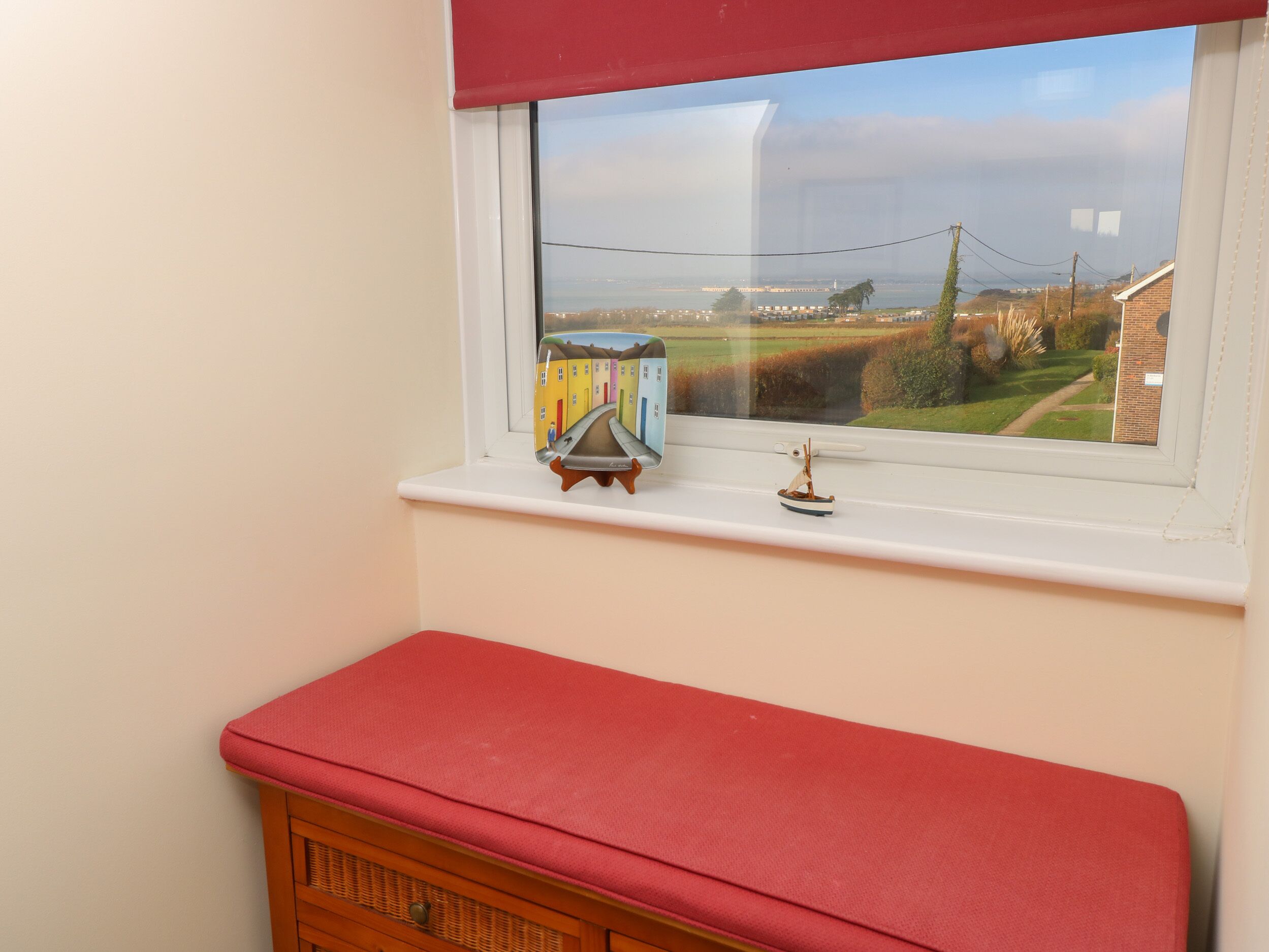 A window with a view of a rural landscape, a small shelf with a model ship, and a red bench below.