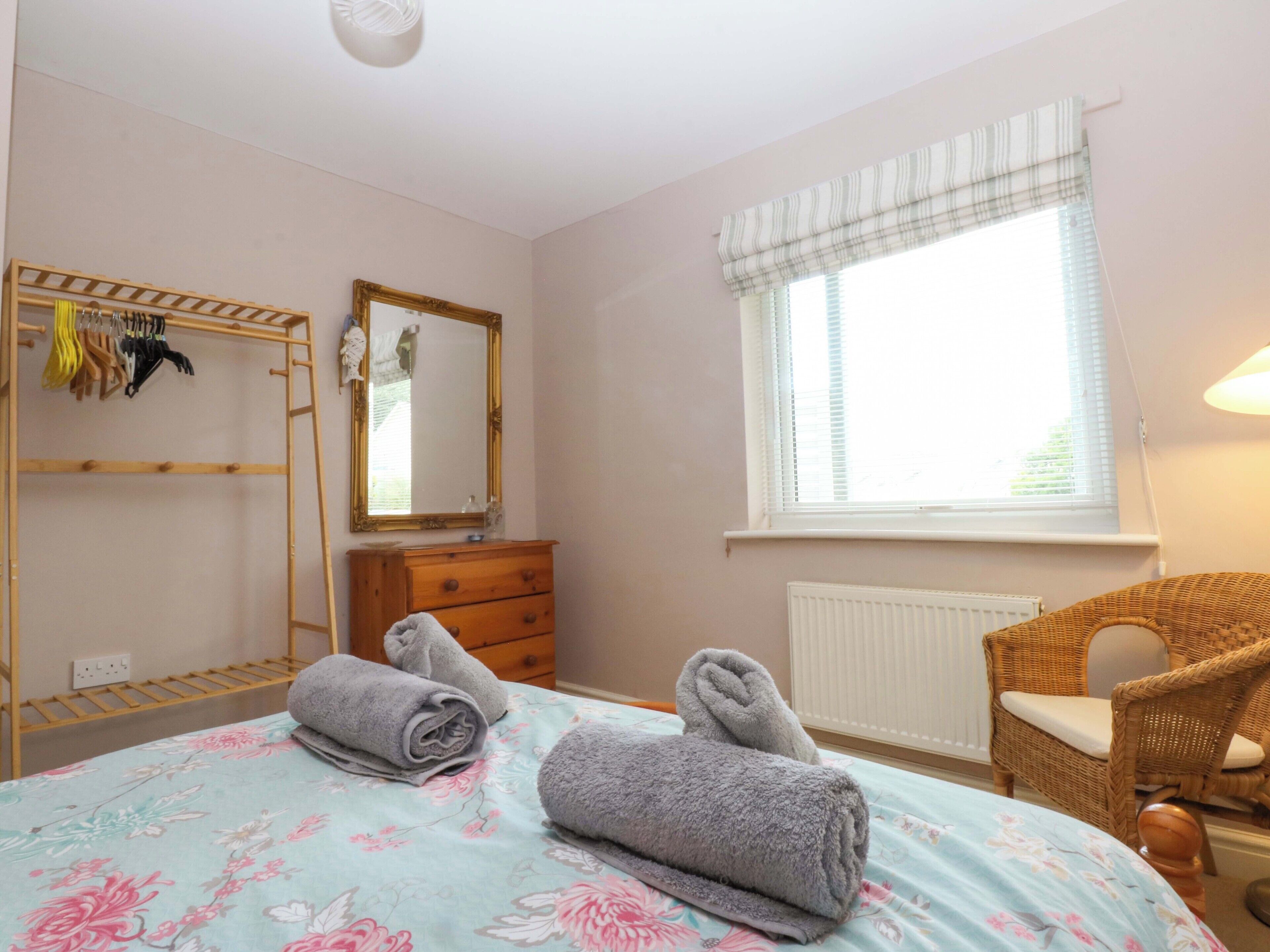 Ein Schlafzimmer mit einem Bett, einem Schminktisch, einem Spiegel, einem Fenster mit Jalousien, einem Korbstuhl und einem Kleiderständer mit Kleiderbügeln.