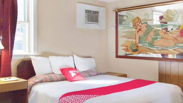A hotel room with a bed, a window with curtains, an air conditioning unit, and a wall-mounted painting. - OYO Hotel Wilkes-Barre East (Wilkes-Barre)