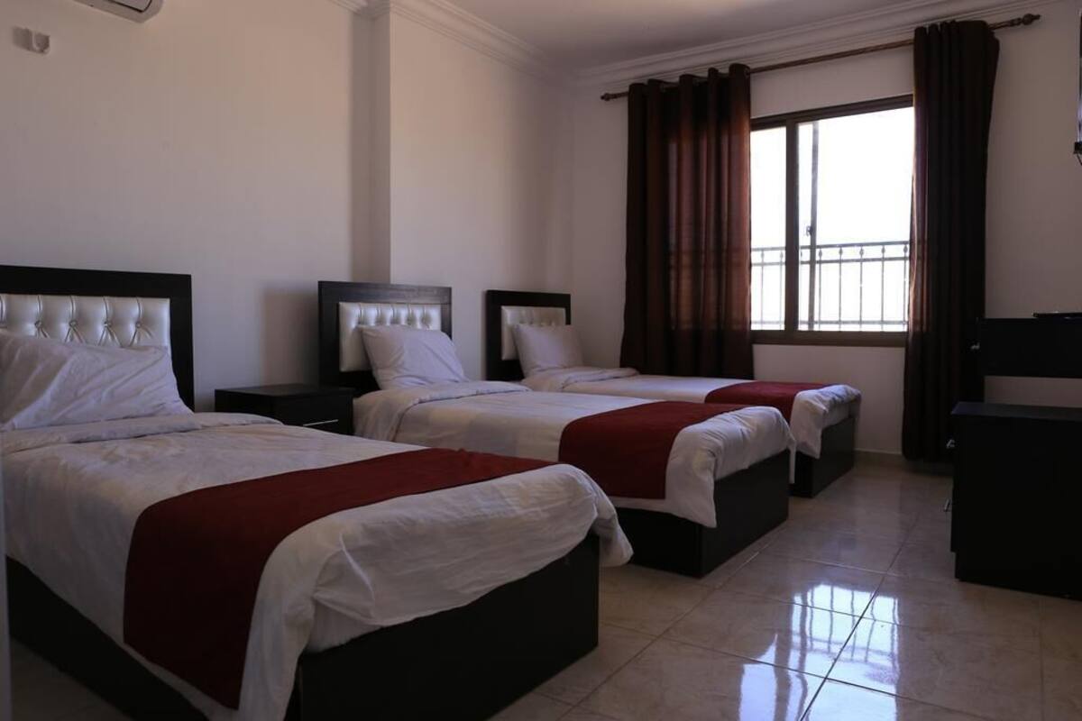 A hotel room with three beds, each with a headboard and white bedding. There is a window with curtains, a small table, and a wardrobe.
