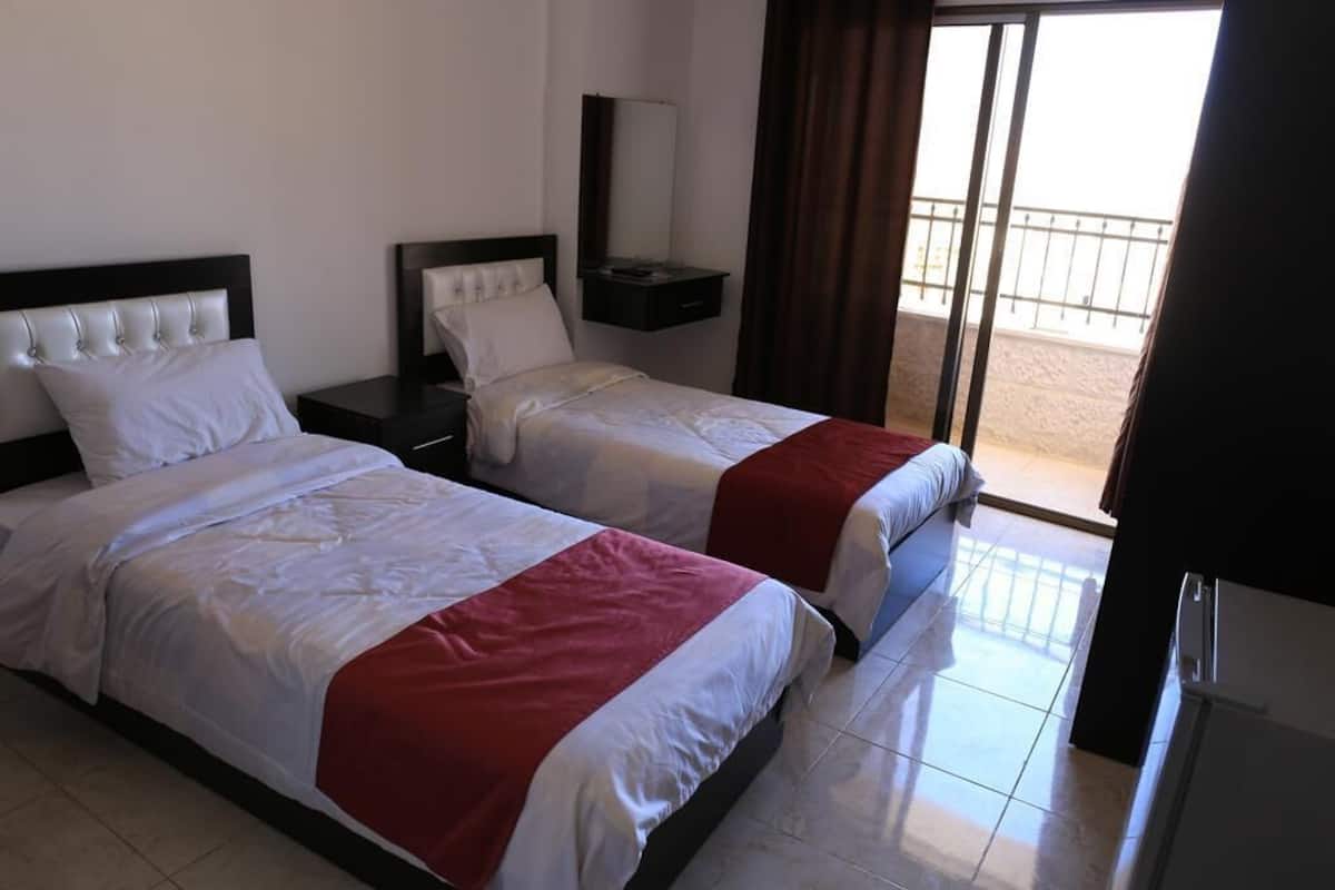 A hotel room with two beds, a small table, a mirror, and a sliding glass door to the outside.