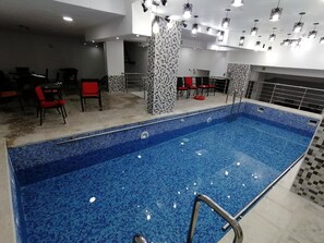 Une piscine aux carreaux de mosaïque bleus, entourée de chaises rouges et noires, et un mur carrelé orné d’un motif géométrique.