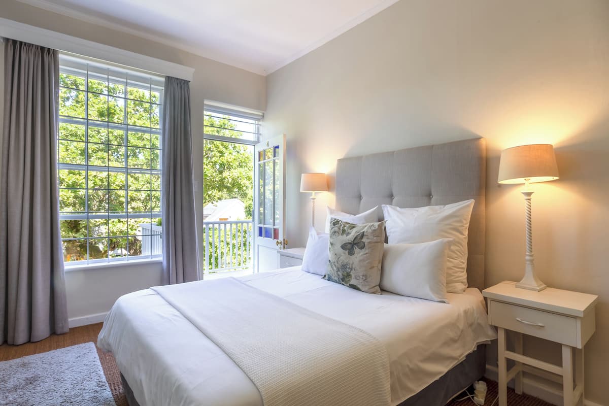 A neatly made bed with white linens and decorative pillows, a bedside table with a lamp, and a window with curtains.