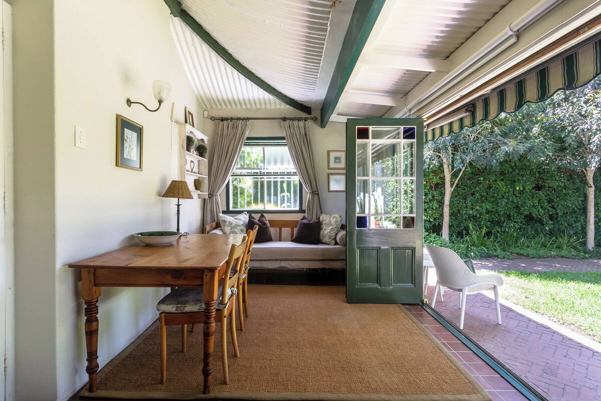 A cozy room with a wooden dining table, chairs, a bench with cushions, a window with curtains, and a view of a garden.