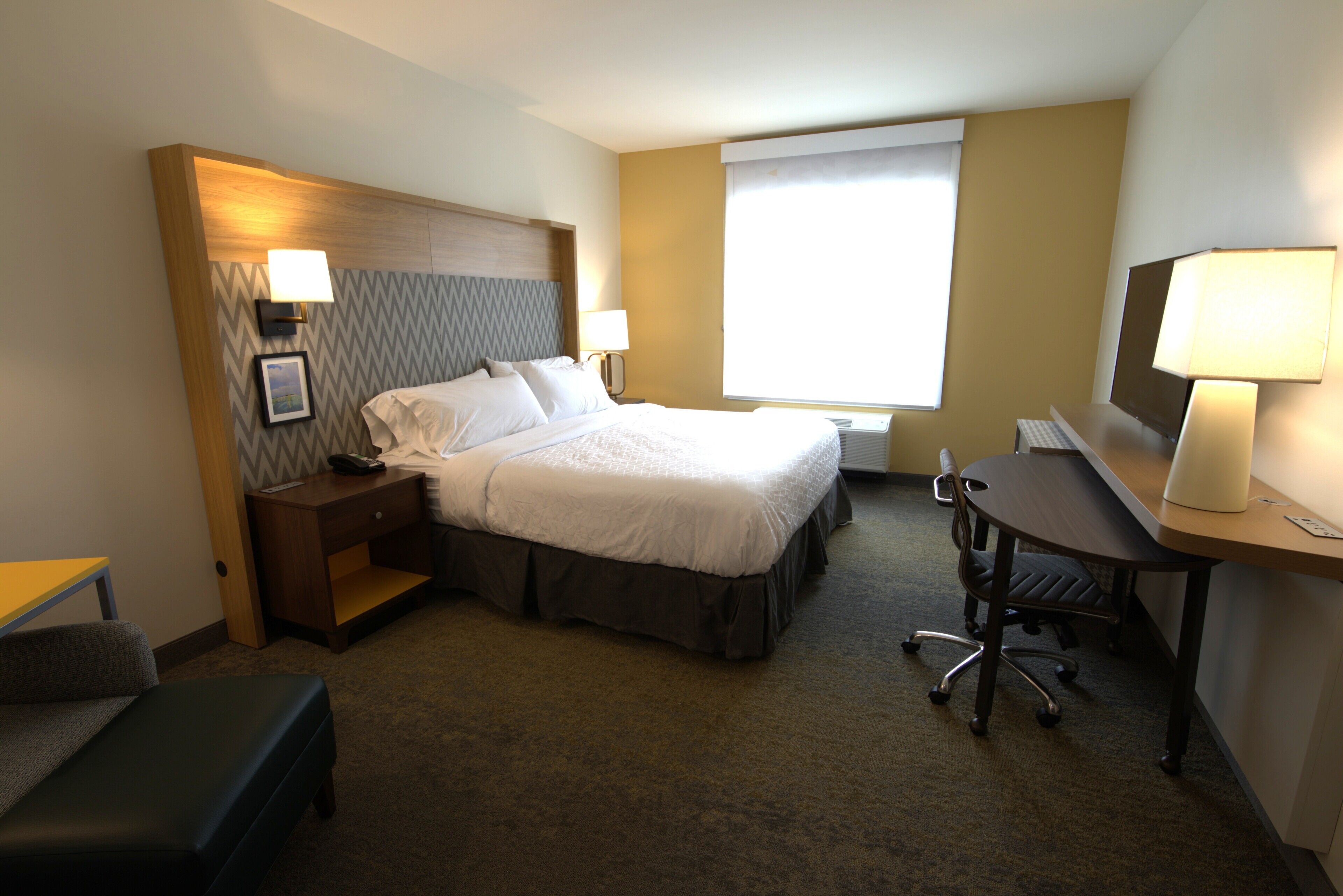 A hotel room with a bed, desk, chair, nightstand, and a large window.