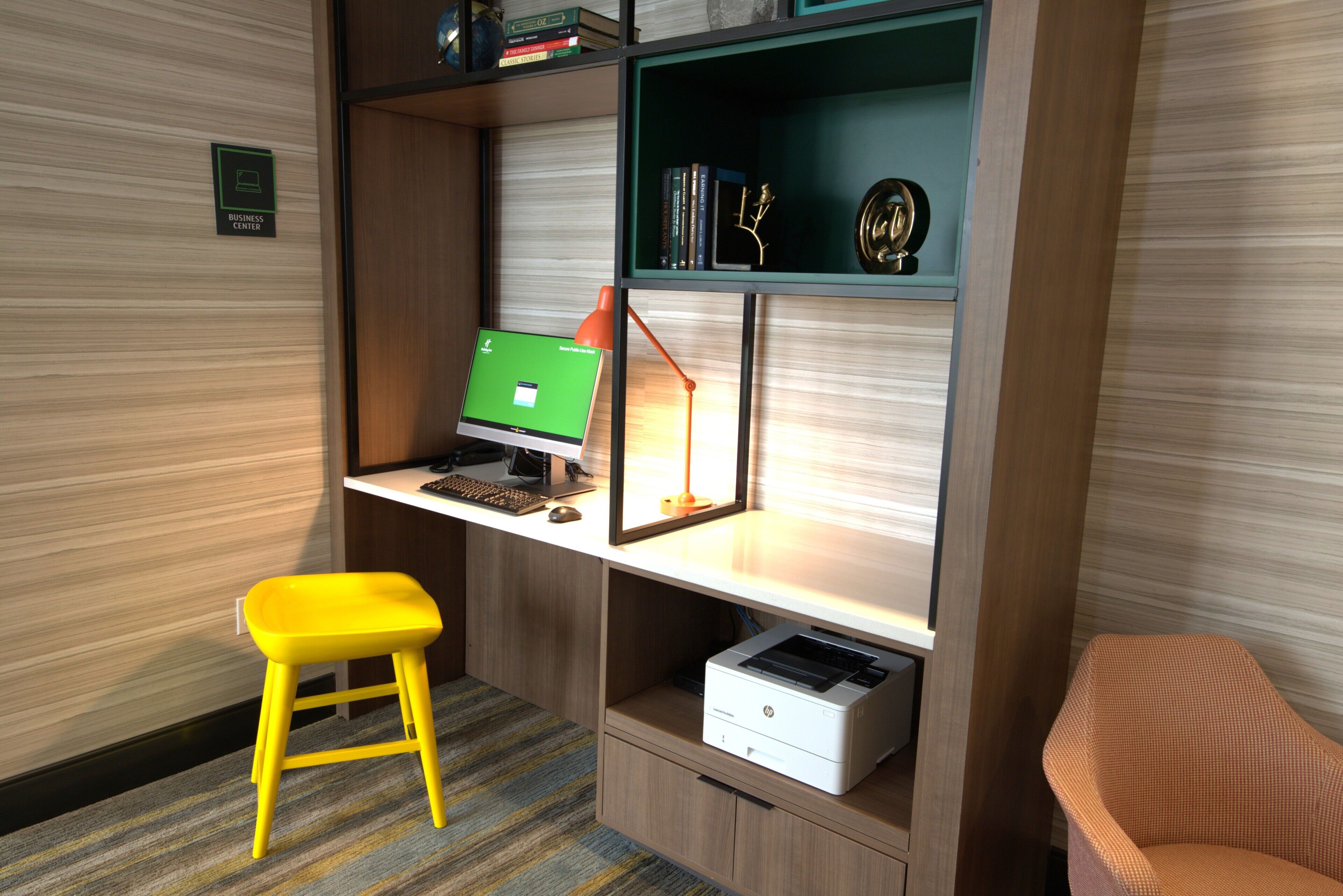 Un espace de travail comprenant un ordinateur, une imprimante, un tabouret jaune et une bibliothèque contenant des livres et des objets décoratifs.