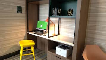 Un espace de travail comprenant un ordinateur, une imprimante, un tabouret jaune et une bibliothèque contenant des livres et des objets décoratifs.