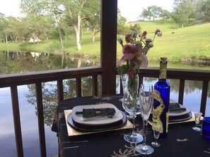 A table set for two with a bottle of champagne, a vase with flowers, and a scenic view of a lake and trees.