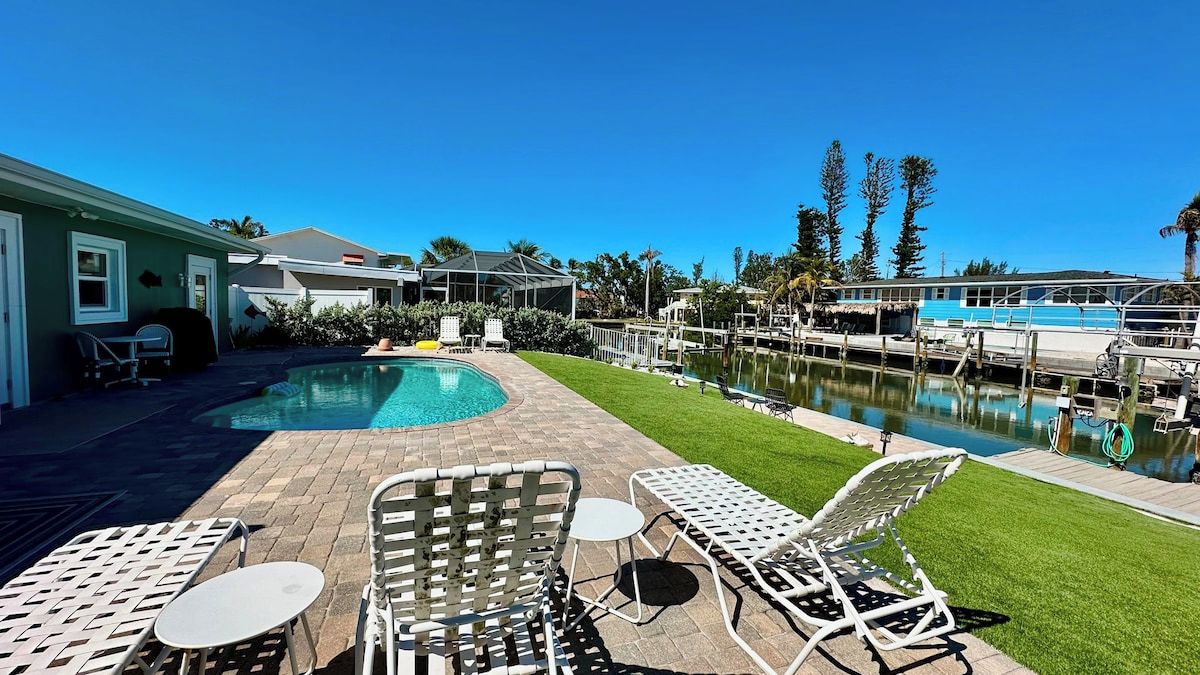 Un jardin avec une piscine, des chaises longues et une jetée qui s’avance sur une étendue d’eau.