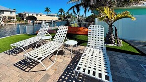 A poolside area with lounge chairs and a small table, surrounded by a fence and palm trees.