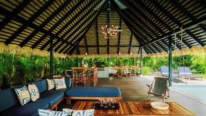 A covered outdoor area with a wooden deck, a sofa with patterned cushions, a dining table, and a pool in the background.