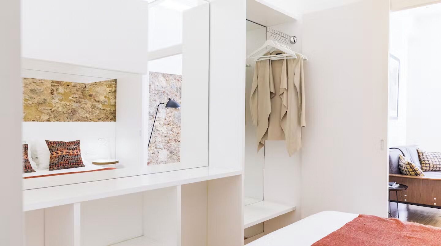 A modern bedroom with a large mirror, a bed with a red blanket, a hanging coat rack, and a stone wall.