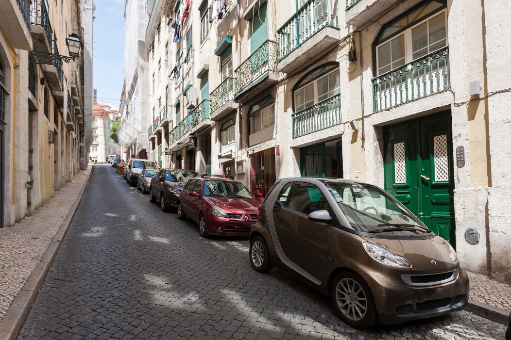 A narrow cobblestone street lined with multi-story buildings, parked cars, and green doors.