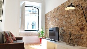 A modern living room with a stone wall, a flat-screen TV, a sofa with patterned cushions, a potted plant, and a floor lamp. - Apt Traditional Charm & Modern Design in Baixa - Lisbon Historic Center (Lisboa)