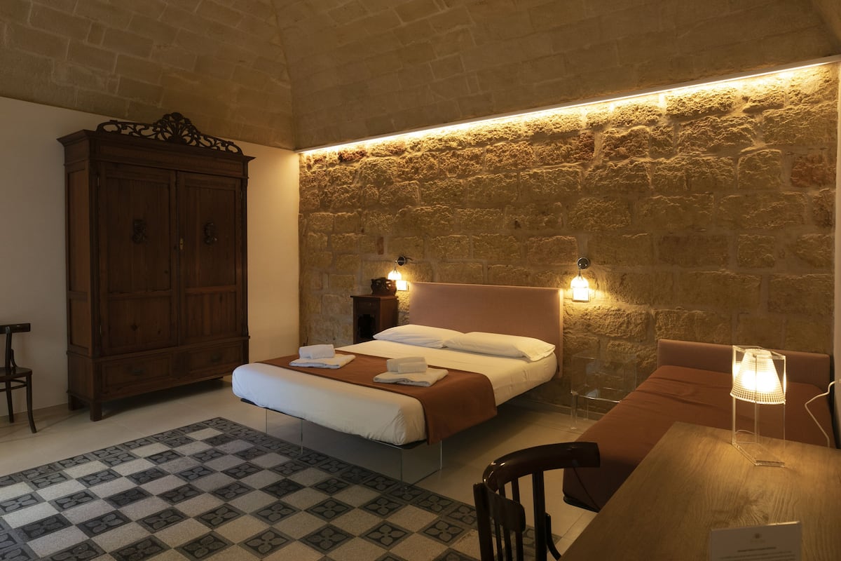 A bedroom with a bed, wooden wardrobe, and a table with a lamp.