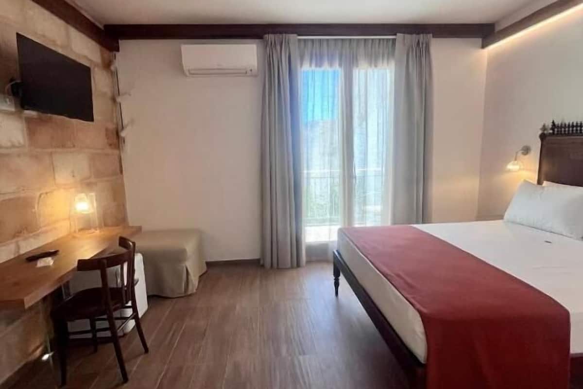A hotel room with a wooden floor, a bed with a red blanket, a desk with a chair, a TV mounted on the wall, and a window with curtains.