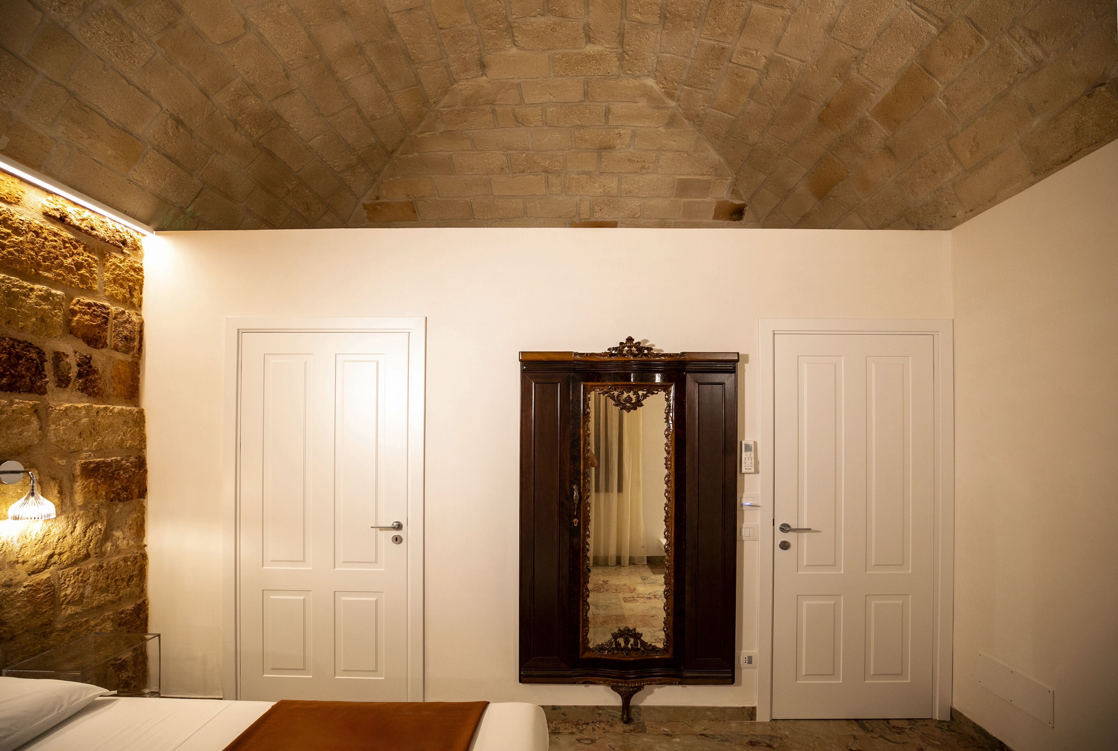 Une chambre avec un mur en pierre, deux portes blanches, un grand miroir et un lit.