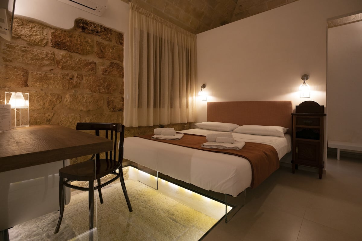 A bedroom with a stone wall, a wooden bed, a nightstand, a chair, and a desk with a lamp.