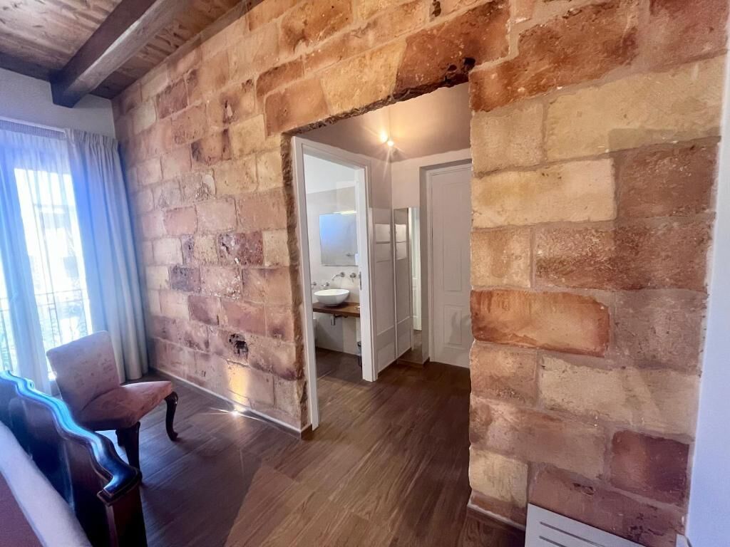 A room with exposed brick walls, a wooden floor, a bench, and a doorway leading to a bathroom with a sink and mirror.