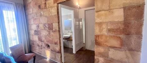 A room with exposed brick walls, a wooden floor, a bench, and a doorway leading to a bathroom with a sink and mirror.