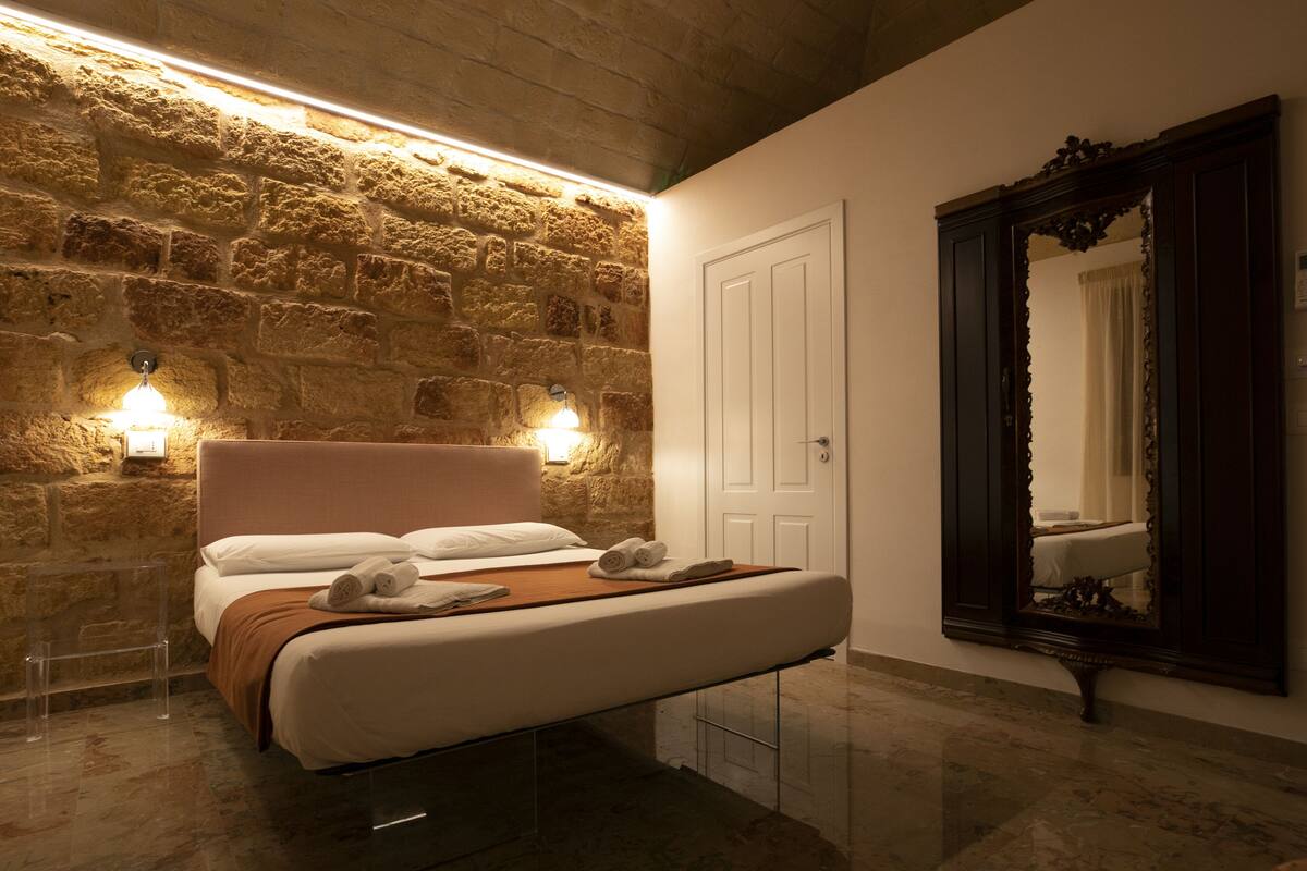 A bedroom with a stone wall, a bed with white linens, two wall-mounted lights, and a large mirror.