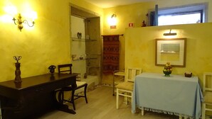 Una habitación con una mesa y sillas de madera, un estante pequeño con adornos, y una ventana con luz natural.