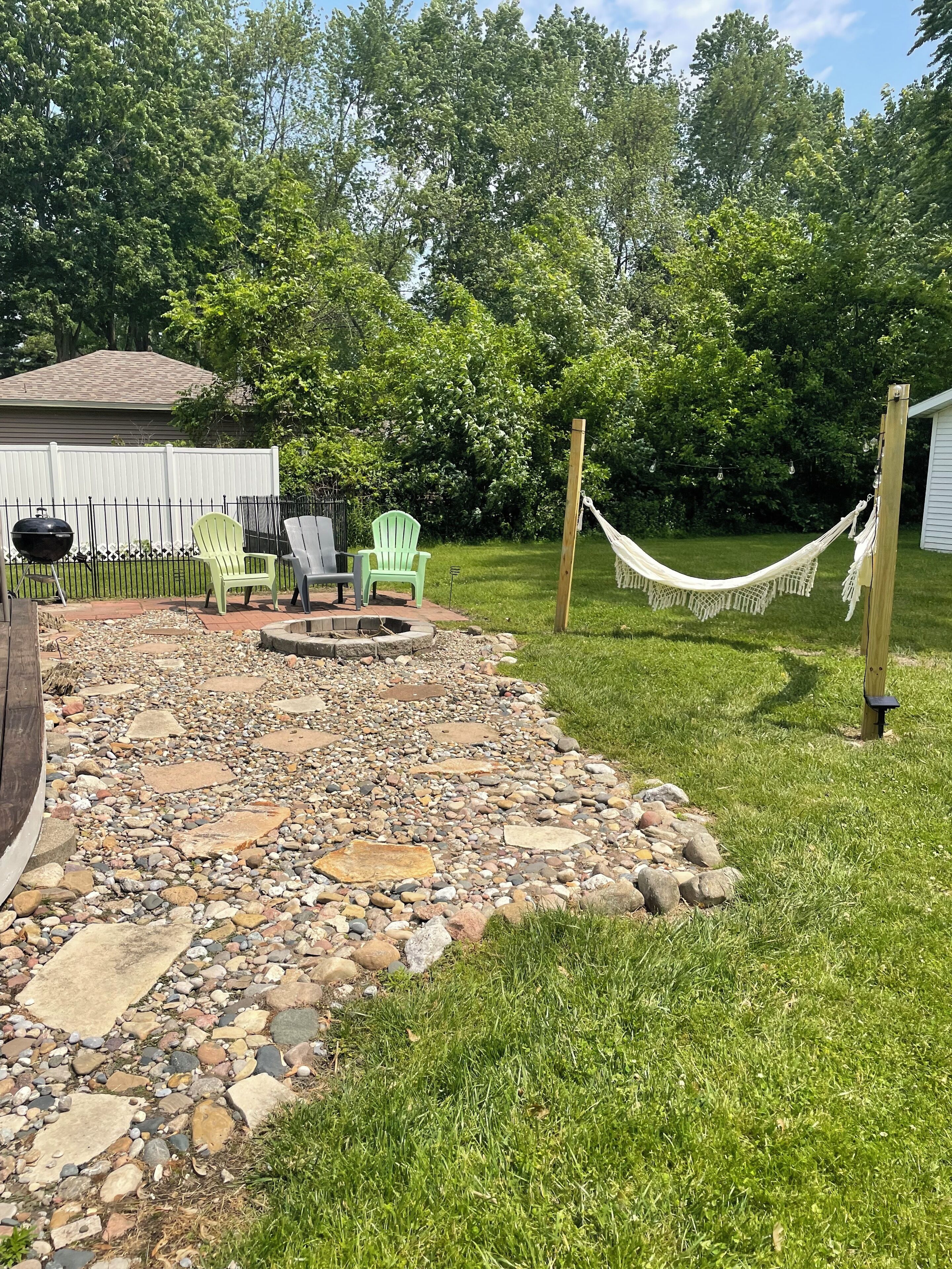 A backyard with a stone patio, two Adirondack chairs, a hammock, and a wooden deck.