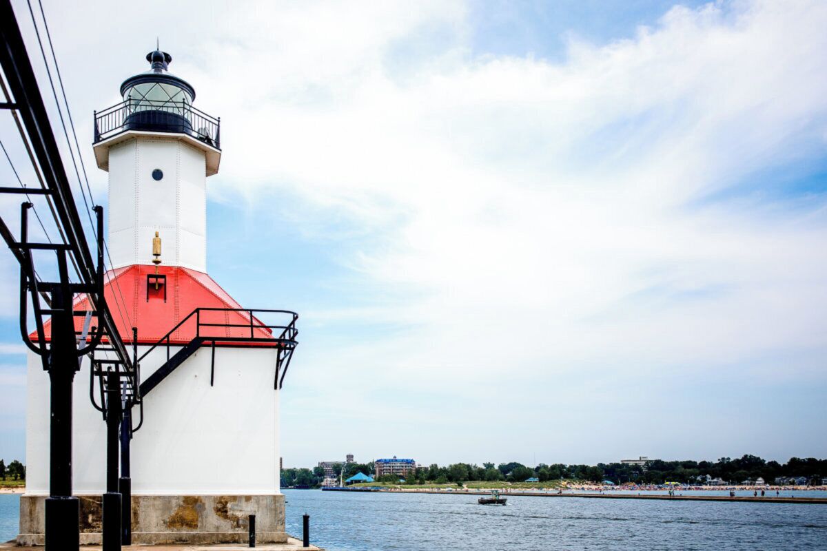 A lighthouse with a red roof, situated by a body of water with a beach and buildings in the background.