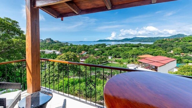 A balcony with a wooden ceiling, a glass table, and a metal railing overlooking a scenic landscape with greenery and a body of water. - Renovated Oceanview Condo w/ Spacious Layout, Covered Terrace & Swimming Pool Near Beach (Playa Flamingo)