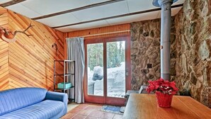 A cozy room with a blue sofa, a wooden table with a red vase, a stone wall, and a large window with a view of a snowy landscape.