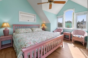 Un dormitorio con cama rosa, dos mesitas de noche con lámparas, una ventana con persianas, un ventilador de techo y vistas a casas.