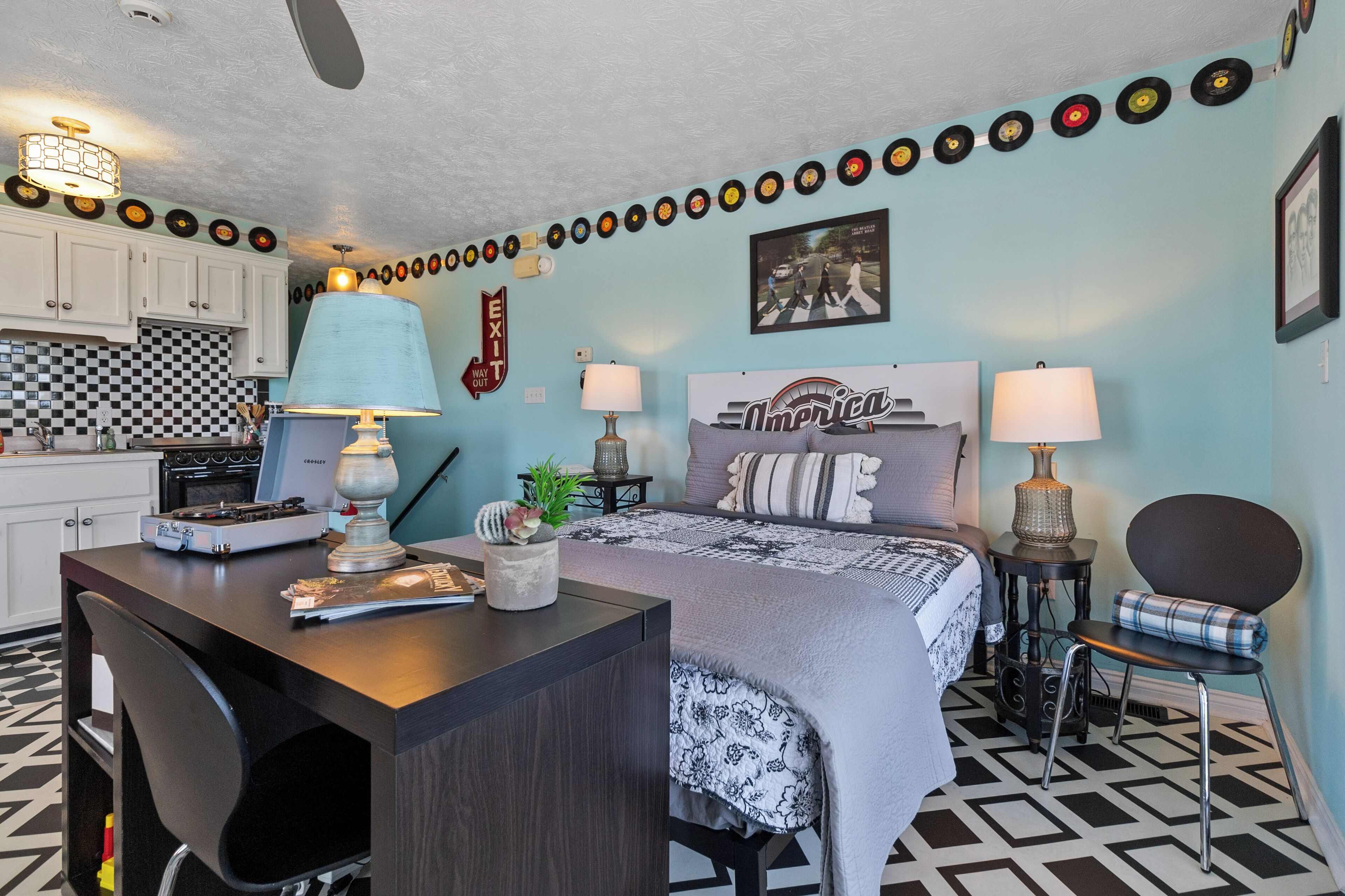 A compact room with a kitchenette, a bed with patterned bedding, and a wall decorated with vinyl records.