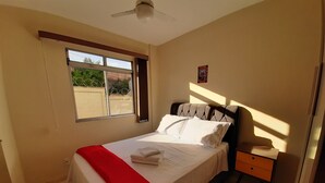 Ein ordentlich bezogenes Bett mit weißen Leinentüchern und einem roten Akzent, ein kleiner Nachttisch mit Lampe, ein Fenster mit Blick auf Grünflächen und ein Deckenventilator.