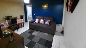 Ein Wohnzimmer mit einer Couch, einem Esstisch und Stühlen. Ein Fenster mit Jalousien, eine Deckenleuchte und ein Wandbild sind ebenfalls vorhanden.