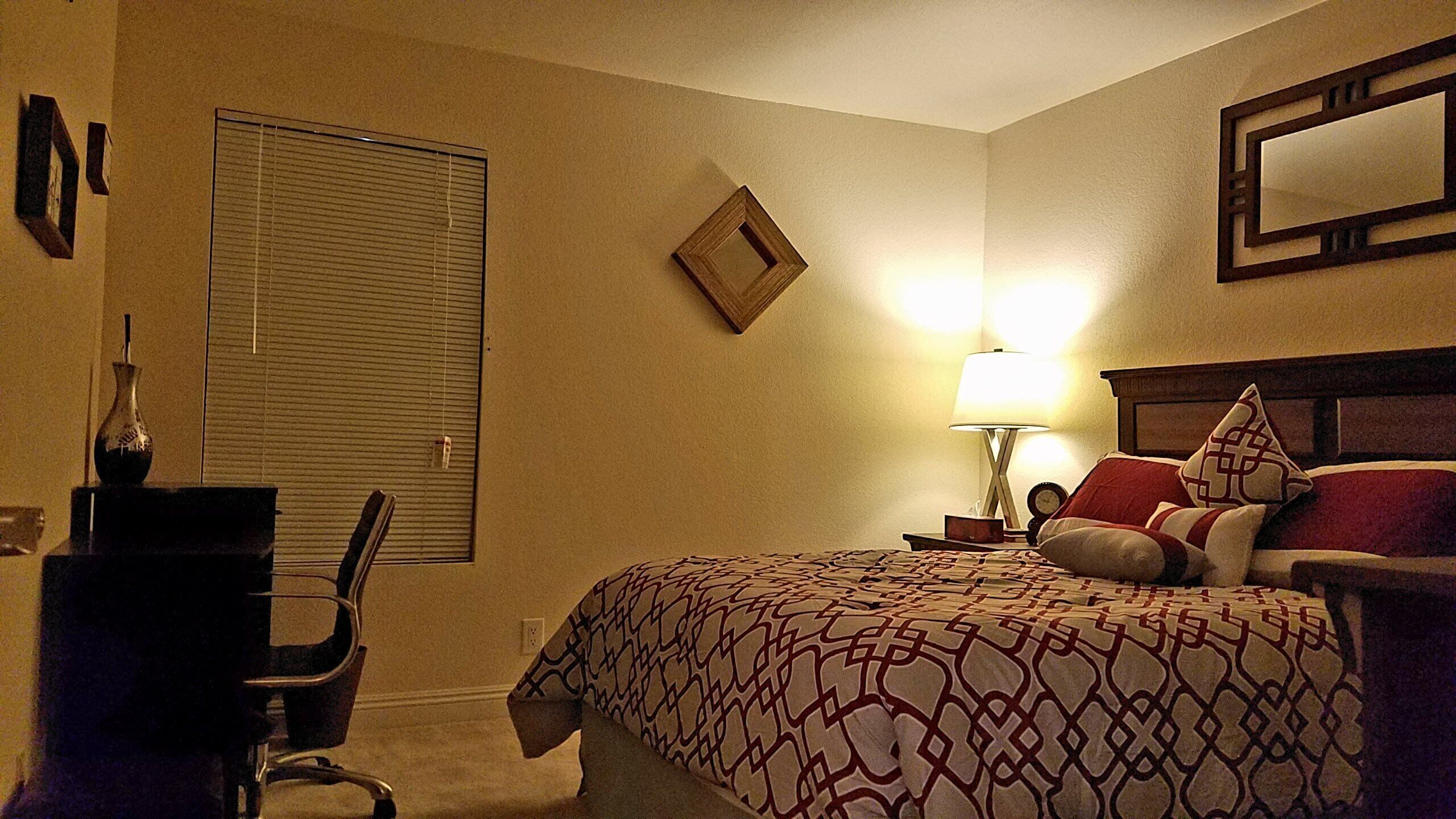 A hotel room with a bed, a desk, a chair, a lamp, and a mirror on the wall.