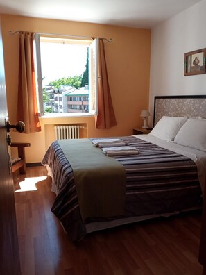 A hotel room with a bed, a window with curtains, a radiator, a bedside table with a lamp, and a painting on the wall.