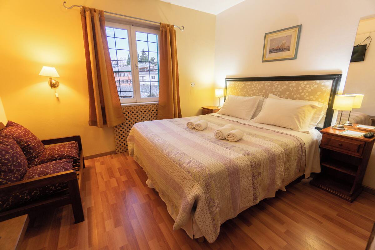 Una cama bien hecha con cabecera, una silla de madera, una mesita de noche con lámpara, y una ventana con cortinas.