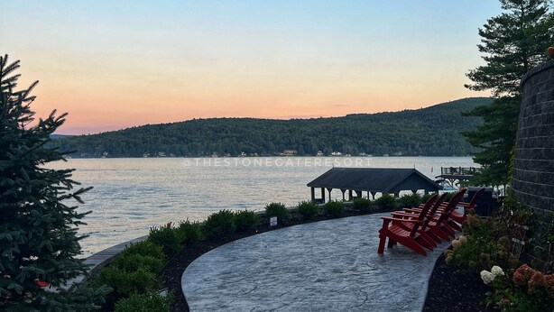 A waterfront patio with a curved pathway, red Adirondack chairs, and a gazebo overlooking a lake.