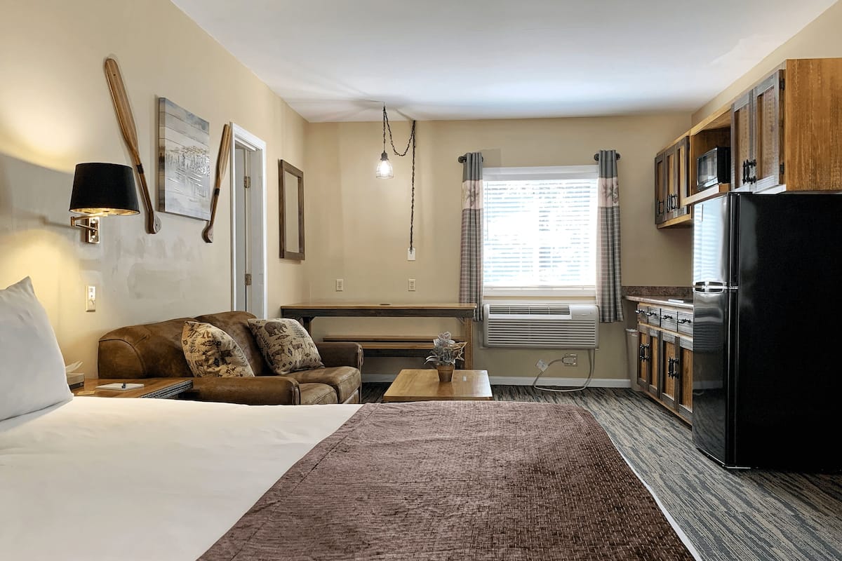 A compact hotel room with a bed, a sofa, a small table, a desk, a kitchen area with a refrigerator, and a window with curtains.