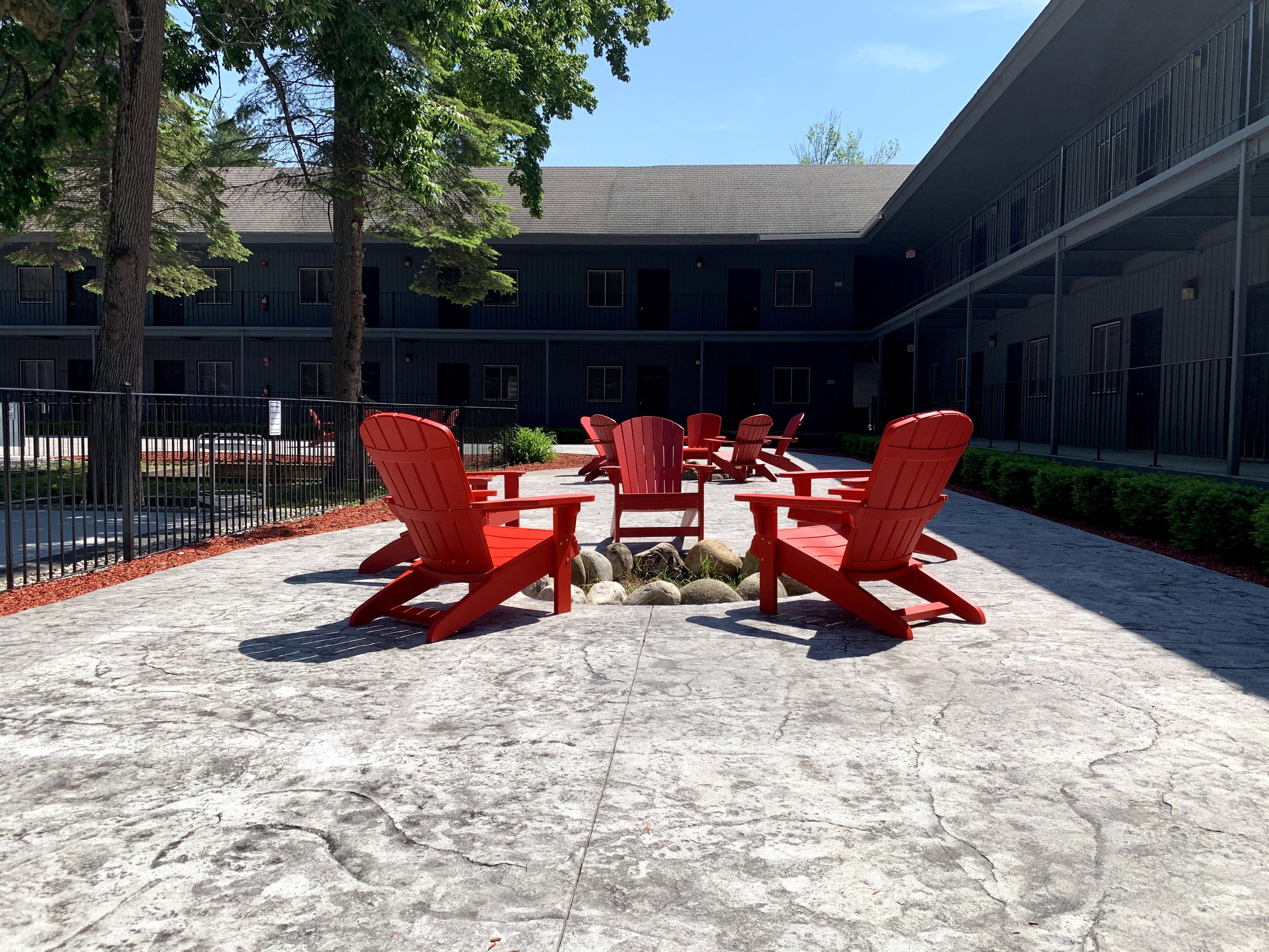 An outdoor seating area with red Adirondack chairs and a concrete table, surrounded by a building with multiple windows and a fenced area.