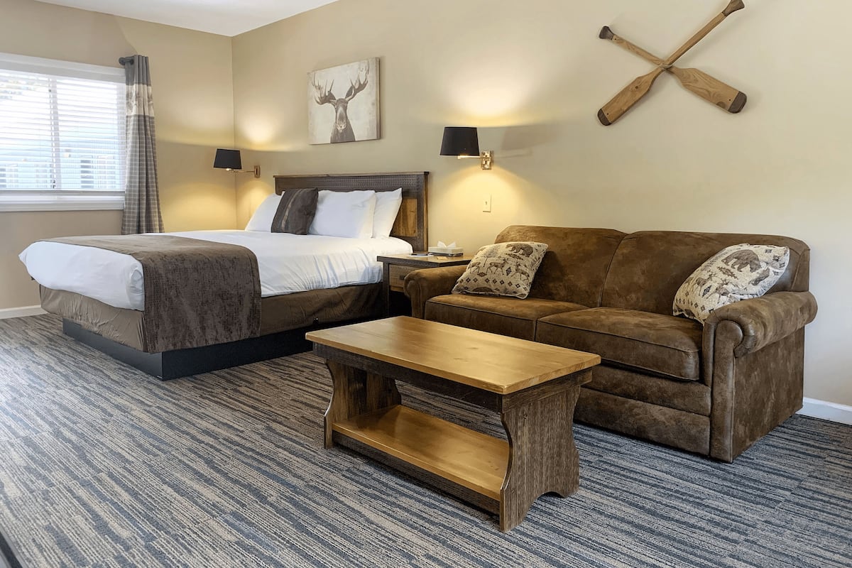 A hotel room with a bed, a brown sofa, a wooden coffee table, and a painting of a moose on the wall.