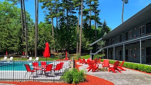 A pool area with red chairs and umbrellas, surrounded by trees and a building with balconies.