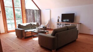 A spacious living room with a large window, a sofa, a coffee table, and a TV on a stand. - Loft "le grenier" (Wolfgantzen)