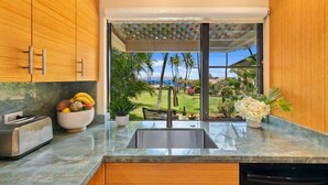 A kitchen with wooden cabinets, a marble countertop, a toaster, a bowl of fruit, and a view of a golf course through a large window. - Wailea Ekahi 34A Spacious Wailea 1BR w AC Pool Near Beach (Kihei)