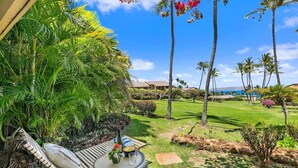 A tropical outdoor seating area with a table, chairs, and a view of a well-maintained lawn and palm trees. - Wailea Ekahi 34A Spacious Wailea 1BR w AC Pool Near Beach (Kihei)